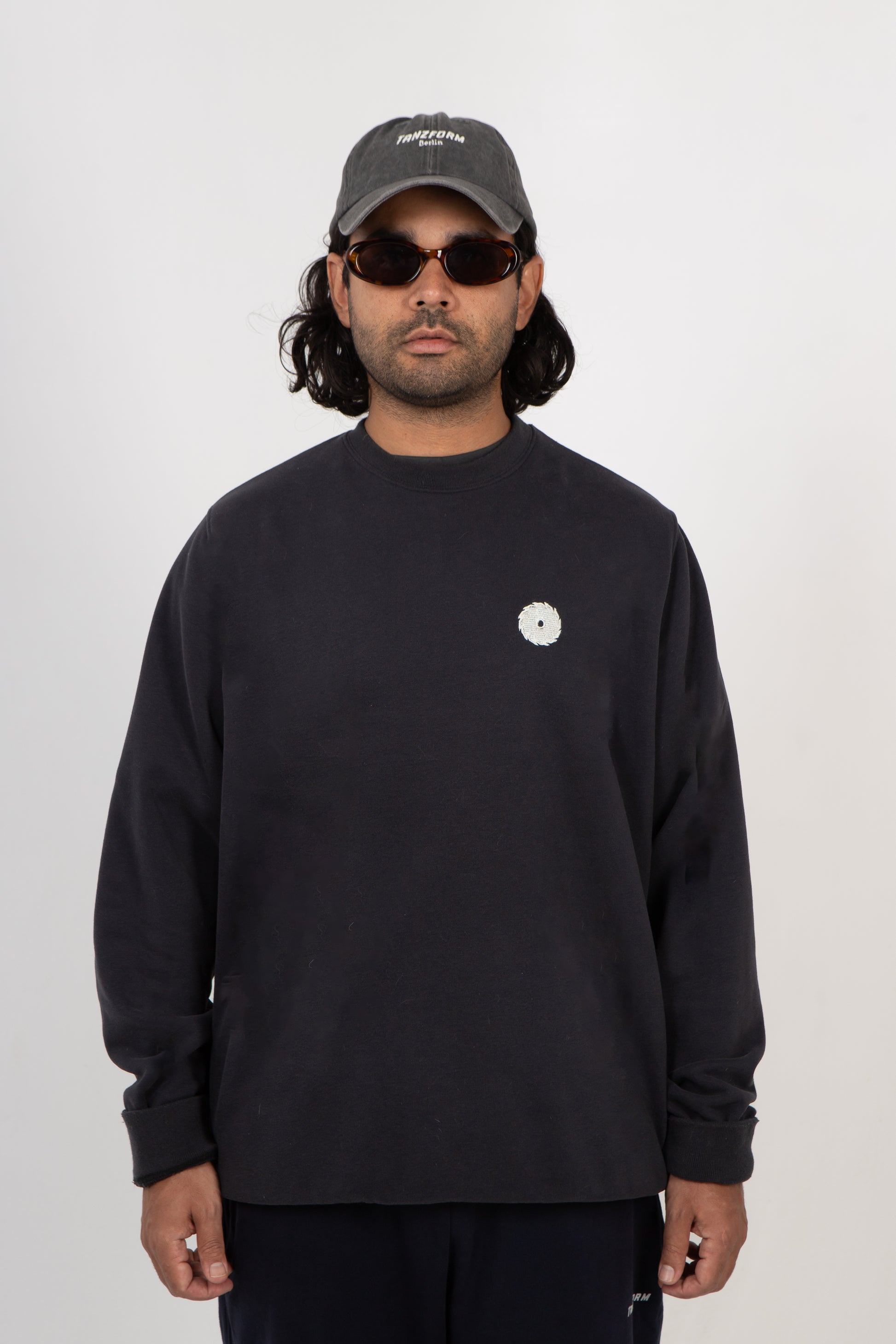 Man wearing a black sweatshirt with a small white logo, black cap, and sunglasses on a white background