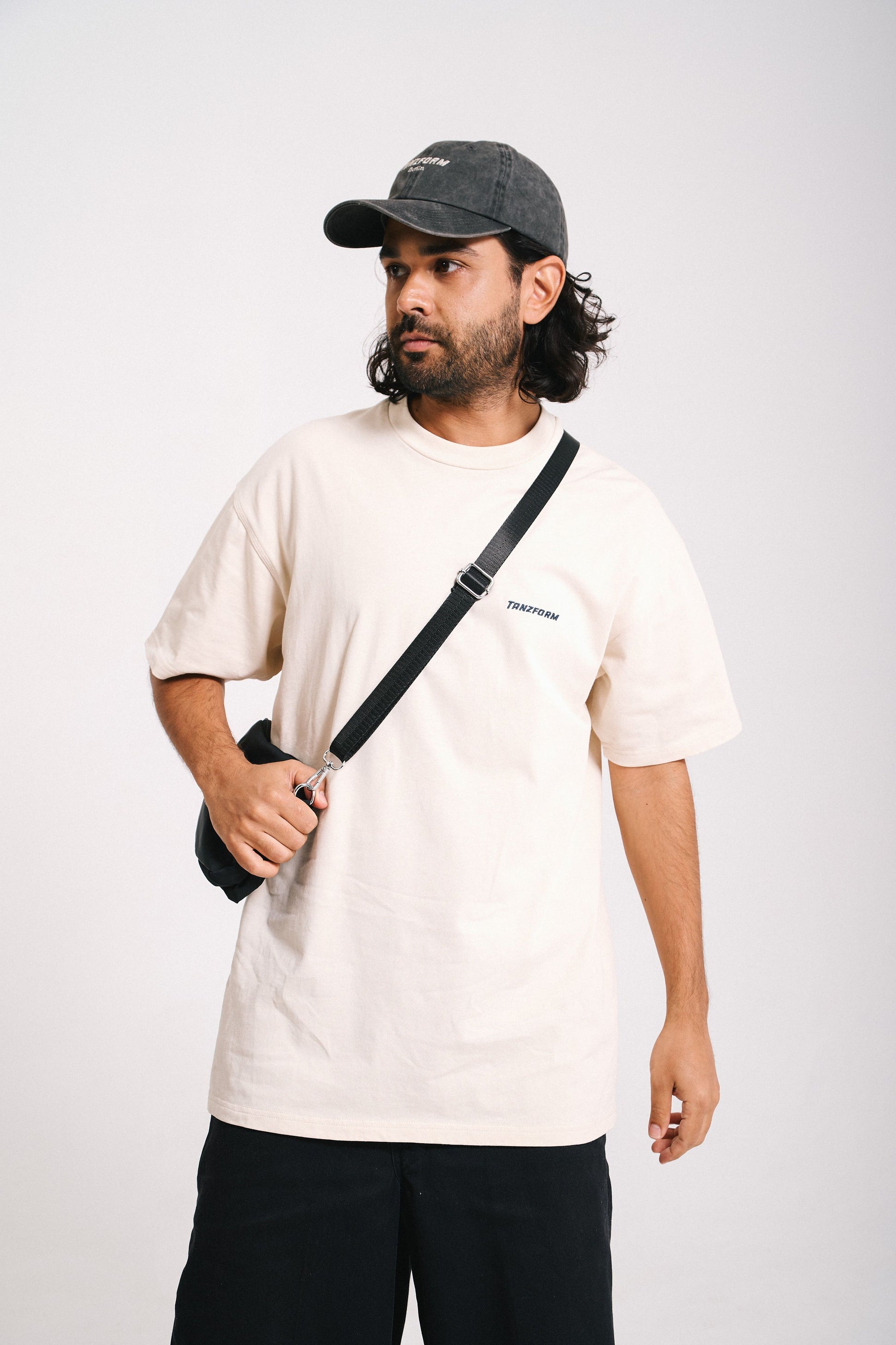 A person wearing a cream white t-shirt and black shorts, holding a camera bag, with a cap on the head.