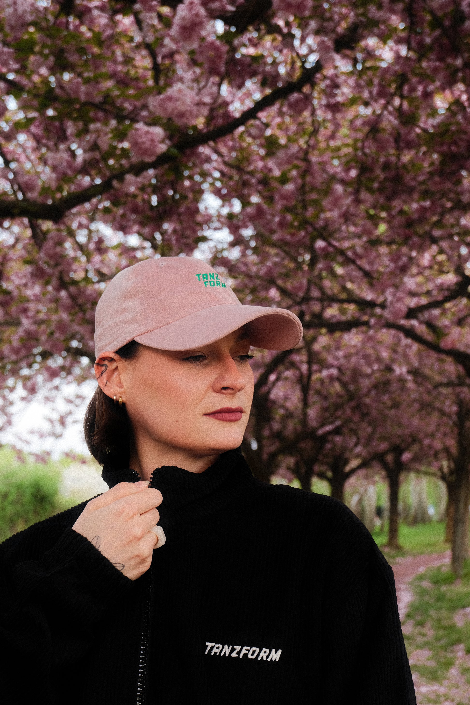 Person wearing a pink cap and black jacket with 'TANZFORM' branding in front of cherry blossom trees.