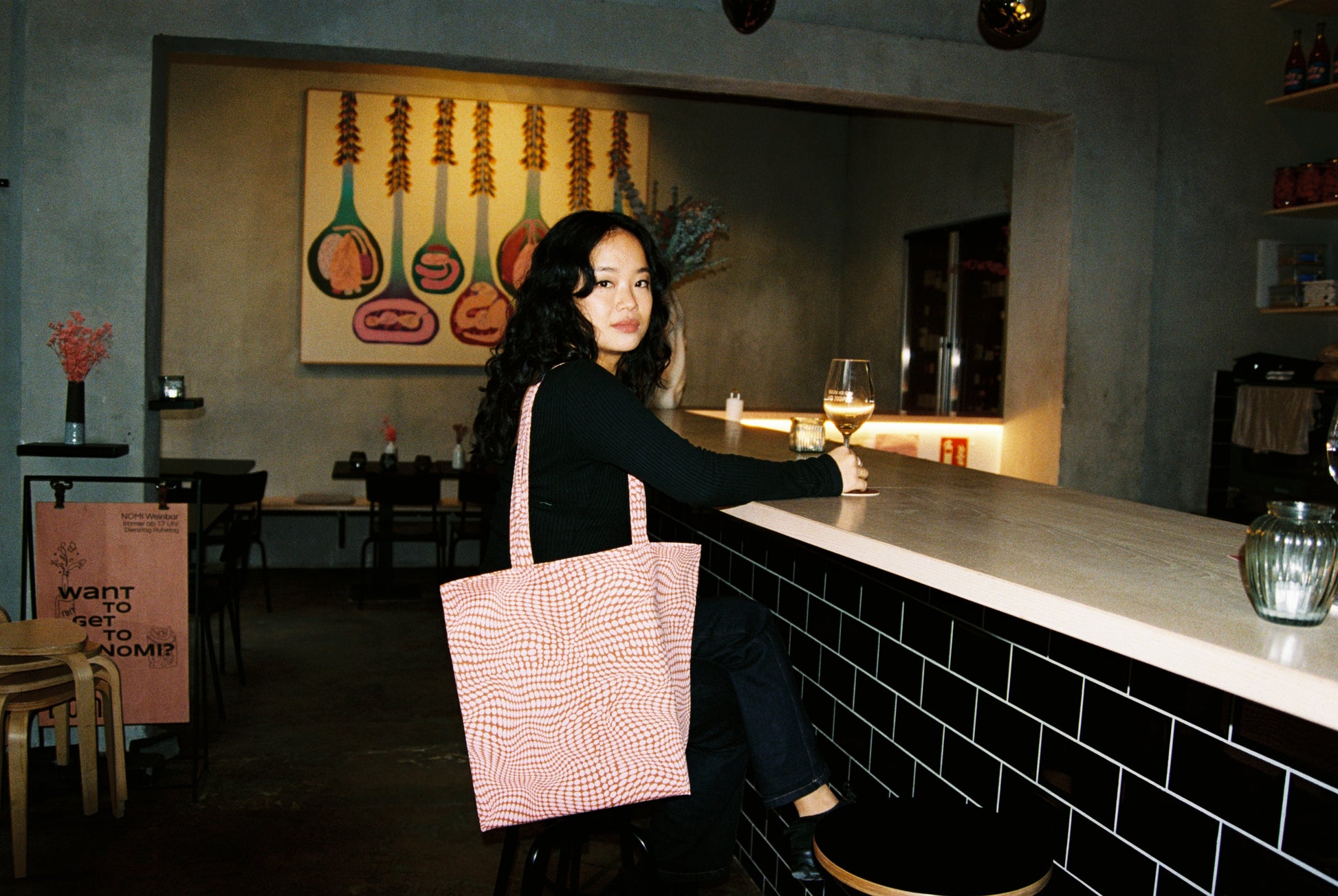 Woman sitting at a bar with a pink bag, artwork on the wall, and a glass of wine.