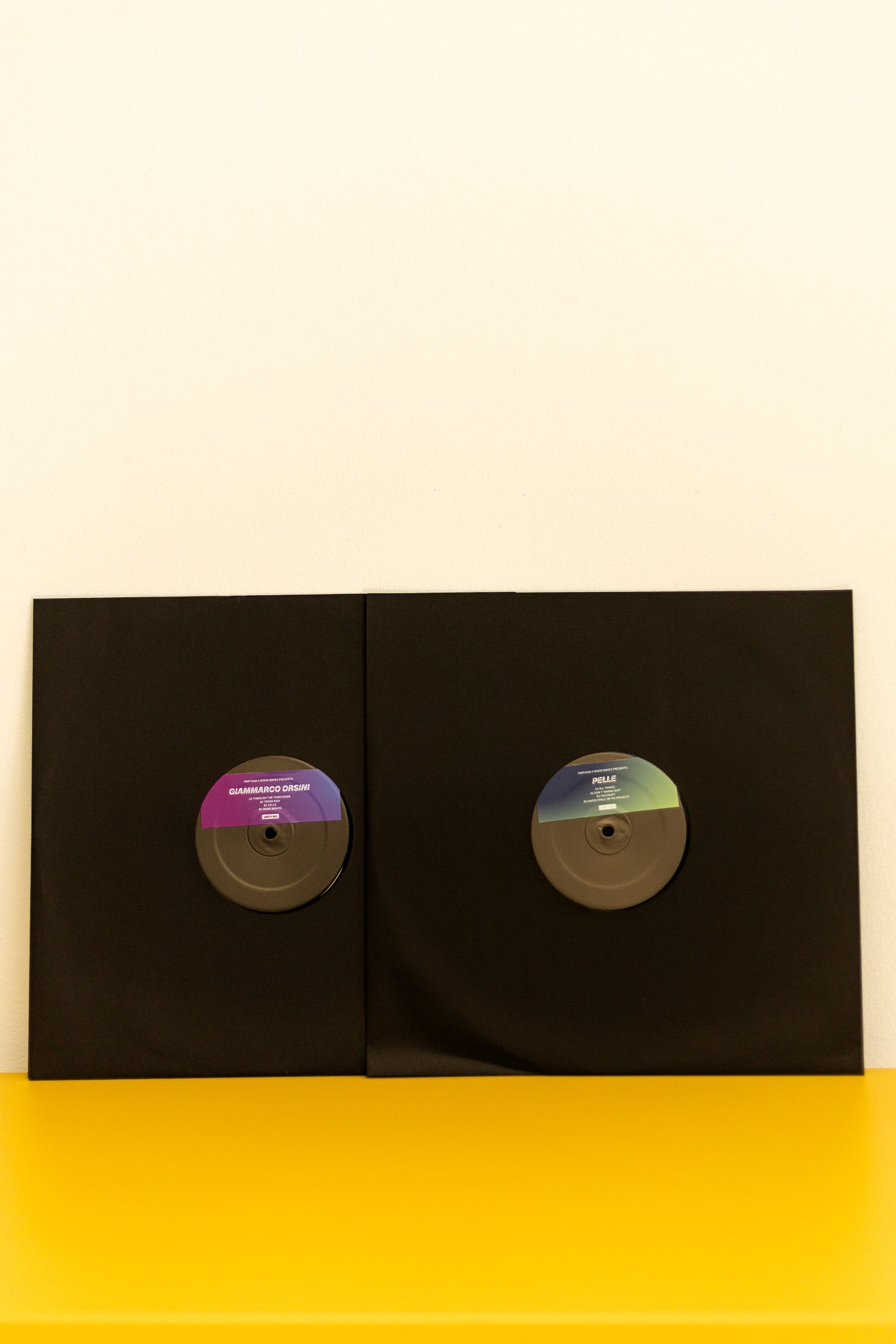 Two vinyl records in black sleeves on a yellow surface with a white background