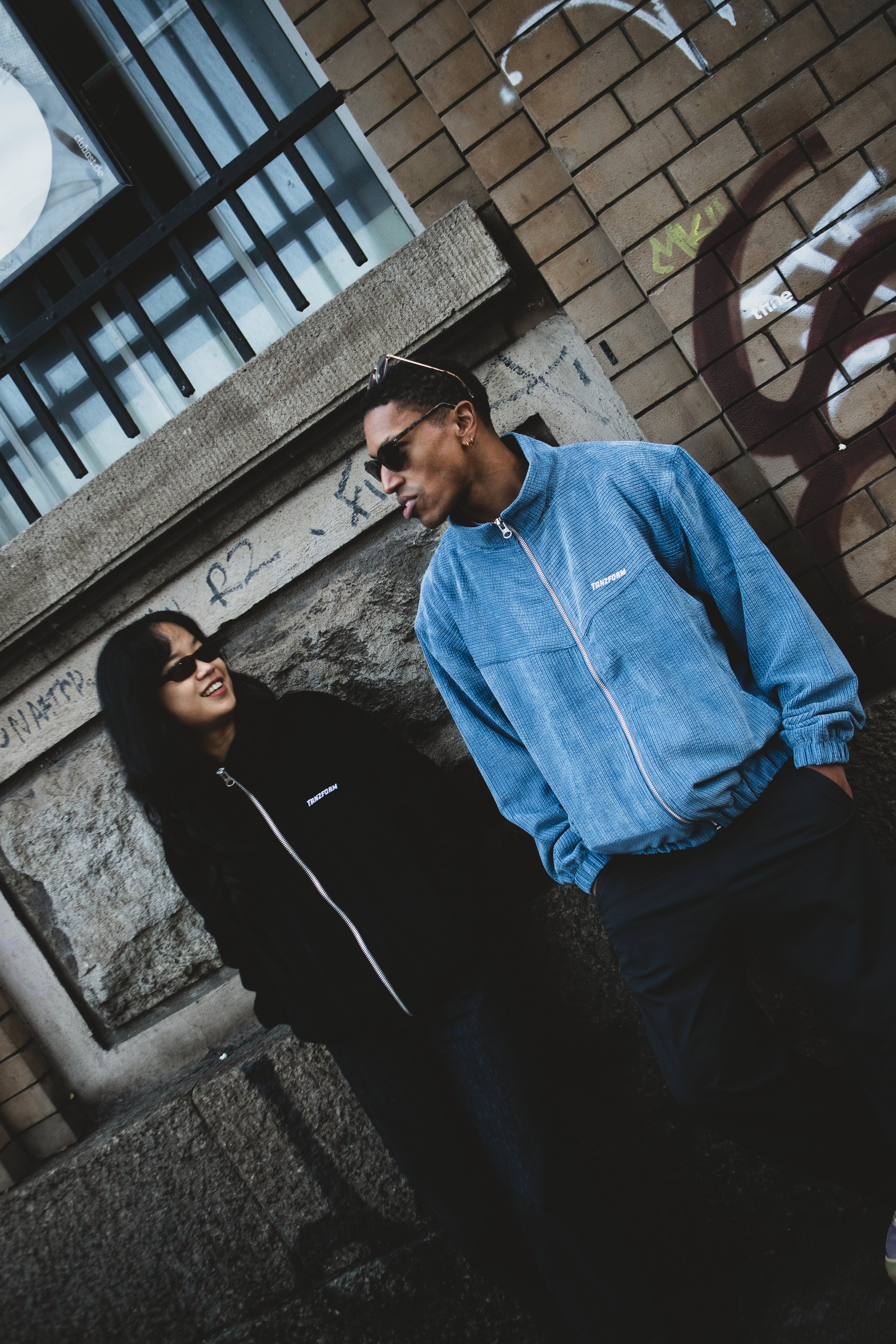Two people standing against a brick wall with a building in the background