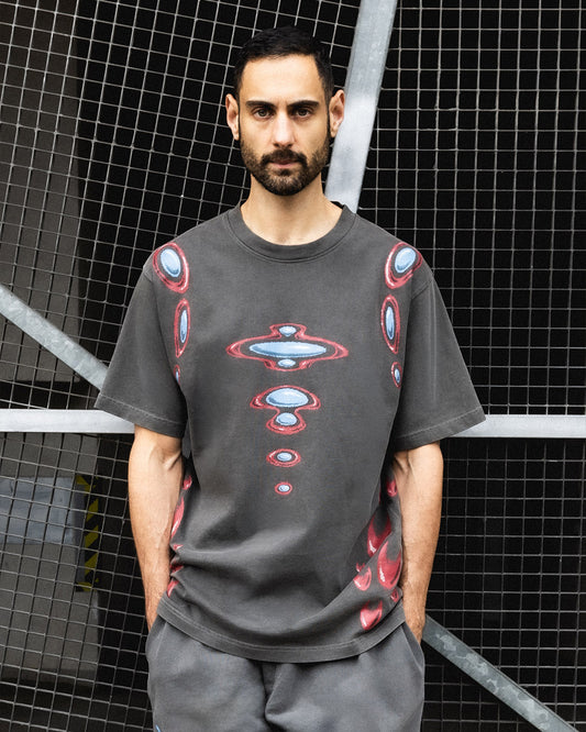 A man wearing a dark gray t-shirt with a red, blue, and white cosmic pattern, standing in front of a black wire mesh grid background.
