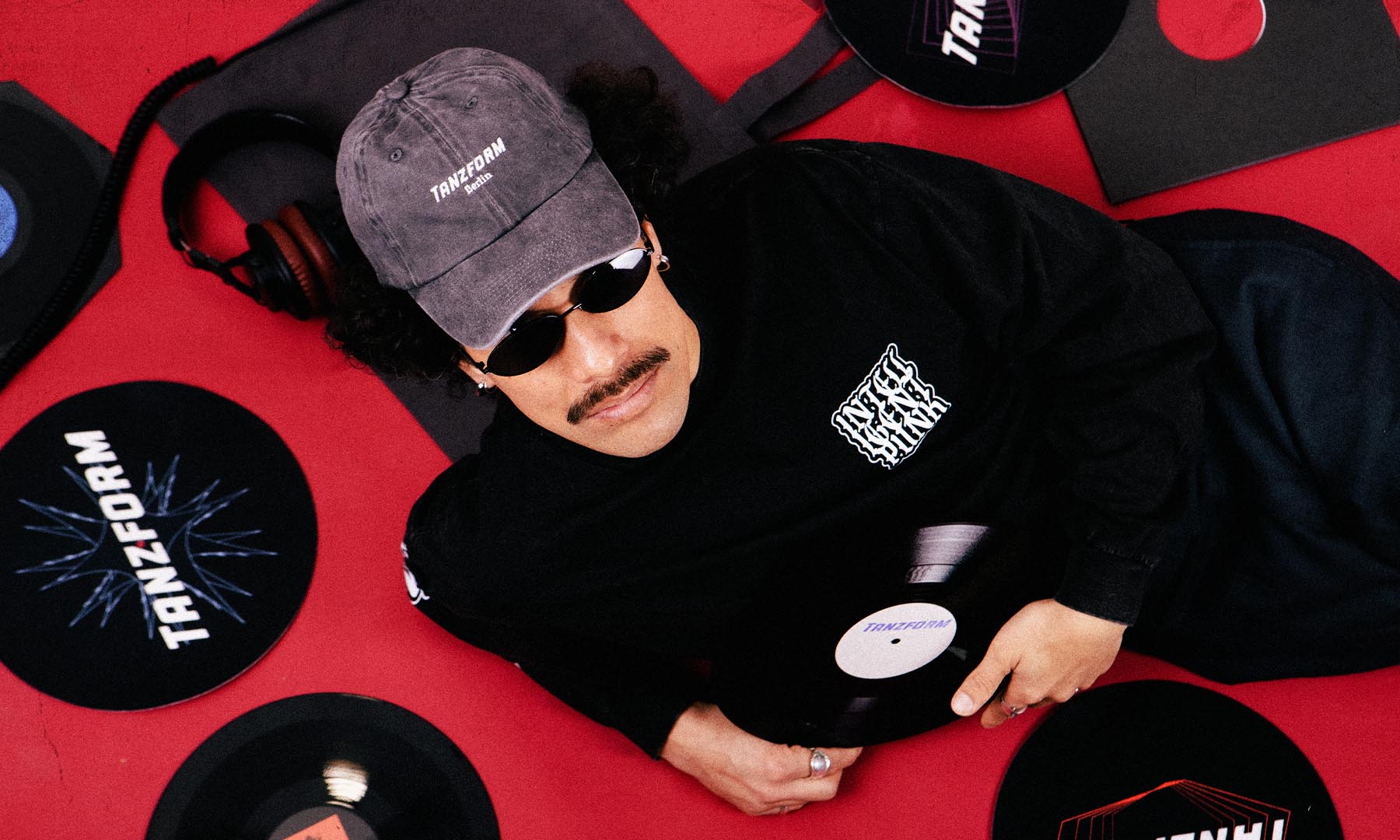 A man wearing a gray cap with 'TANZERM' and black sunglasses lies on a red floor surrounded by black vinyl records and headphones. He is wearing a black shirt with a white graphic logo.