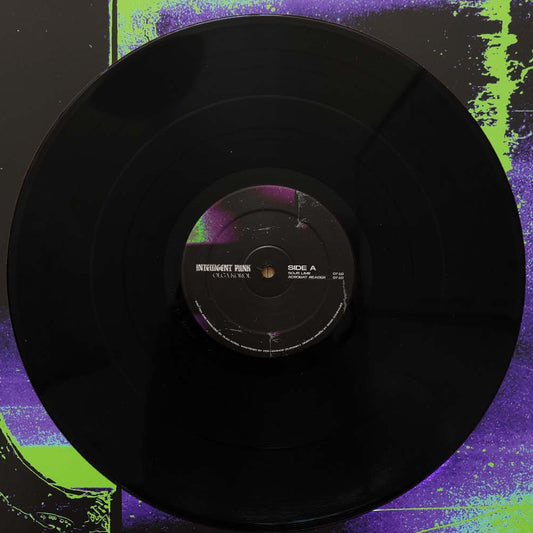A black vinyl record with a purple and white center label, photographed against a dark background with bright neon green and purple paint-like splatters.