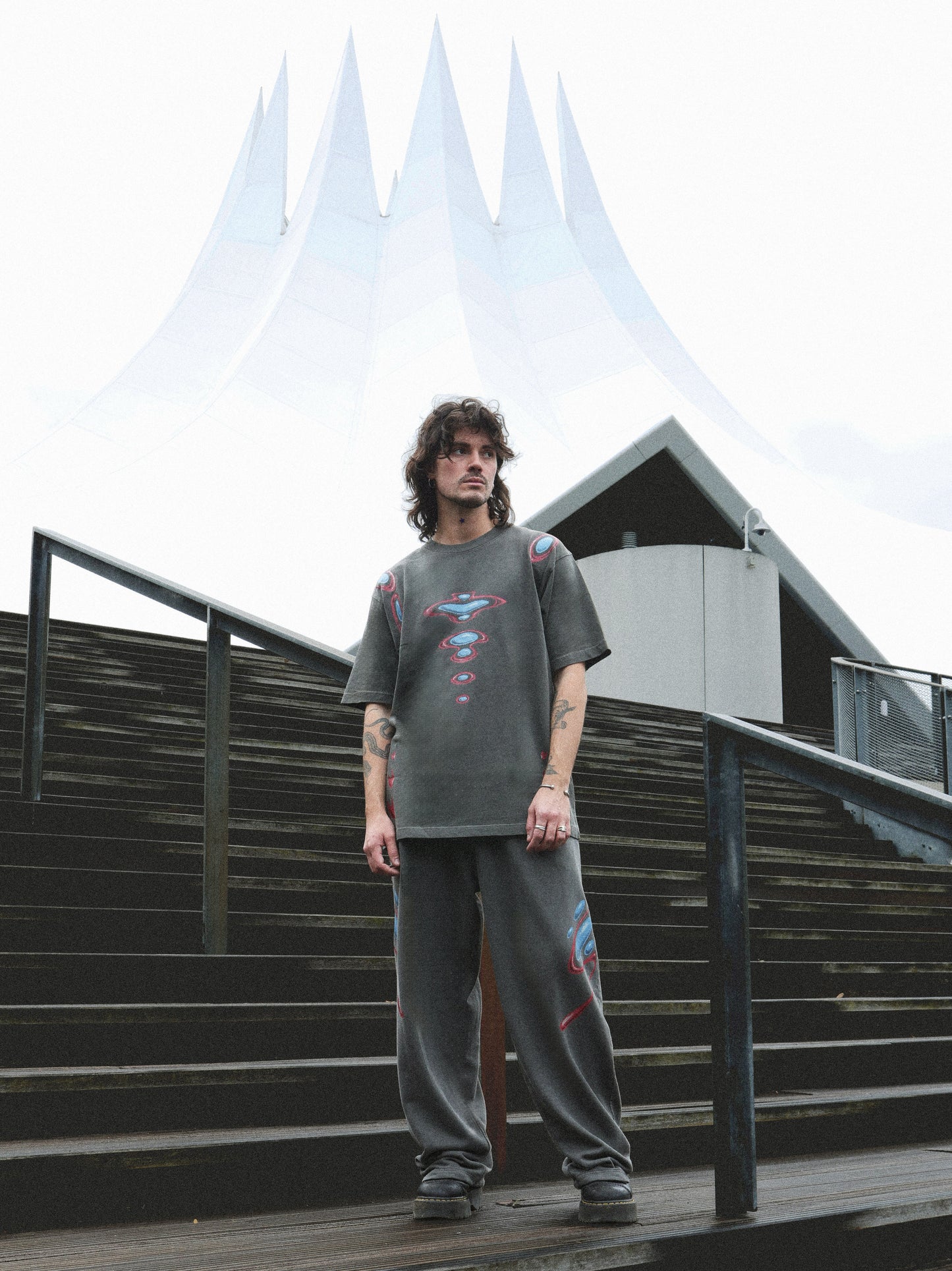 A man wearing a dark gray graphic t-shirt and matching sweatpants stands on outdoor wooden stairs in front of a modern white tent-like building.