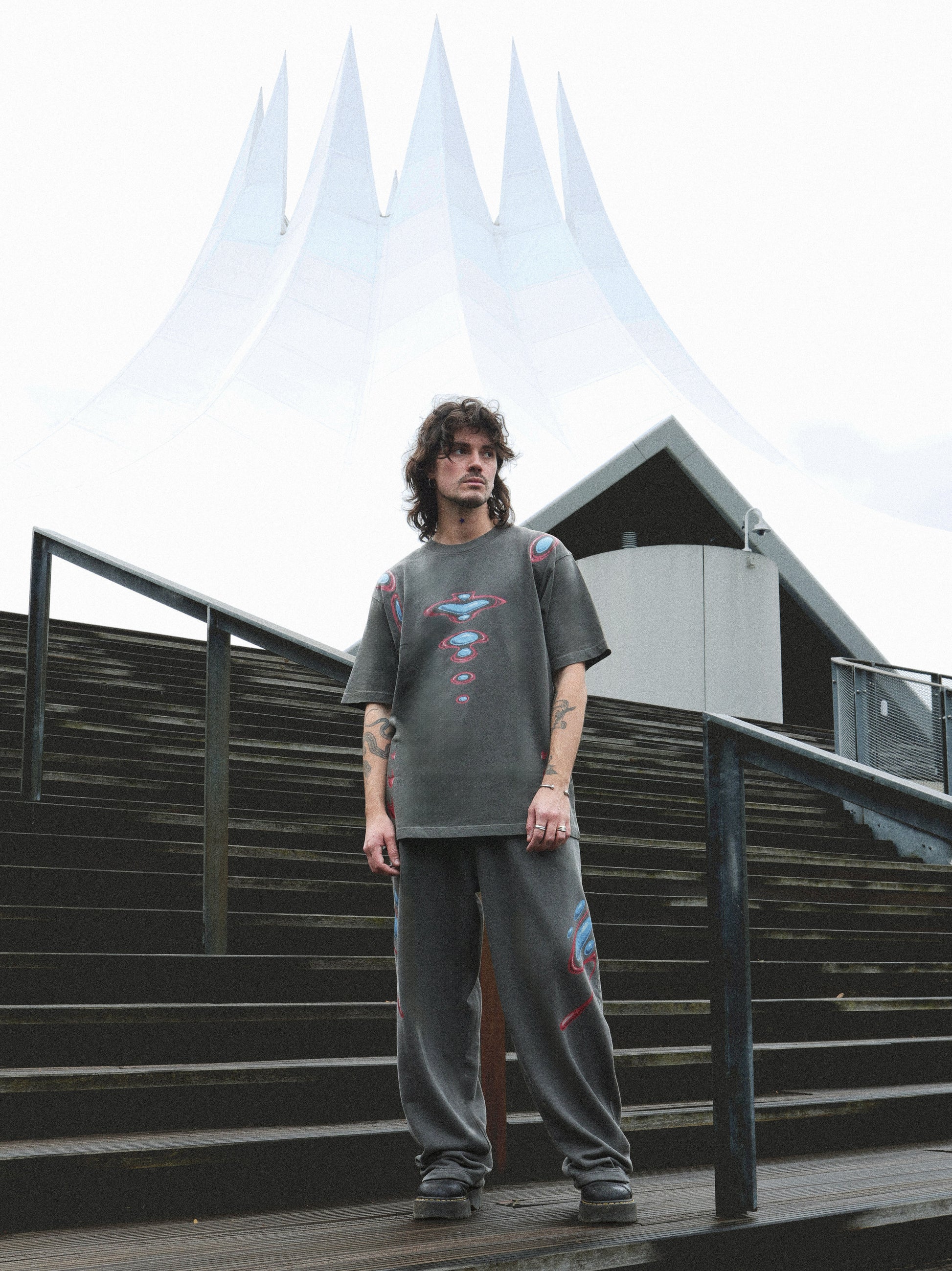 A man wearing a dark gray graphic t-shirt and matching sweatpants stands on outdoor wooden stairs in front of a modern white tent-like building.