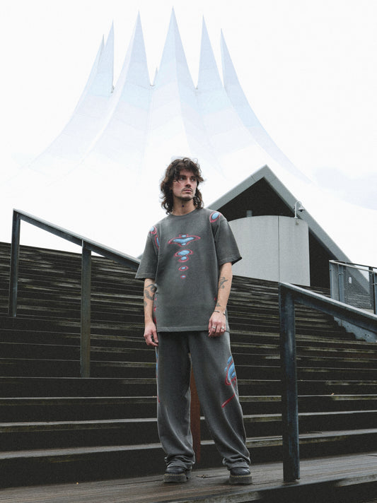 A man wearing a dark gray graphic t-shirt and matching sweatpants stands on outdoor wooden stairs in front of a modern white tent-like building.